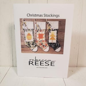 May include: A printable pattern for making three Christmas stockings. The stockings are decorated with a patchwork design featuring a Christmas tree, a star, and a bauble. The pattern is by Rebecca Reese, @17cherrytree_lane.