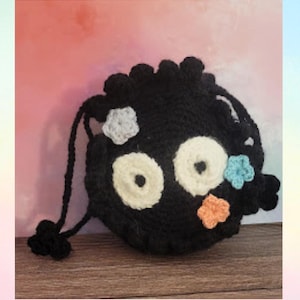 May include: Black crocheted drawstring bag with a fluffy texture and colorful flower embellishments. The bag has two large white eyes and a small black nose.