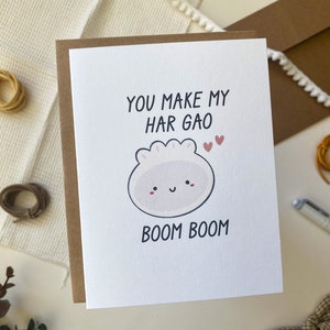 May include: A white card with brown trim features a cartoon illustration of a har gao dumpling with a smiling face. The text on the card reads "You make my har gao boom boom" with two small pink hearts above the dumpling.