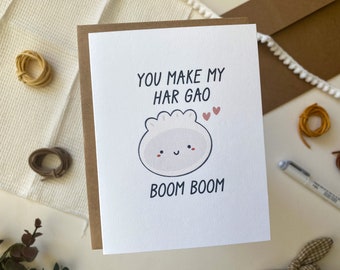 May include: A white card with brown trim features a cartoon illustration of a har gao dumpling with a smiling face. The text on the card reads "You make my har gao boom boom" with two small pink hearts above the dumpling.