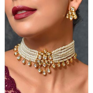 May include: A white pearl choker necklace with a gold and clear stone centerpiece. The necklace has multiple strands of pearls and a teardrop design.
