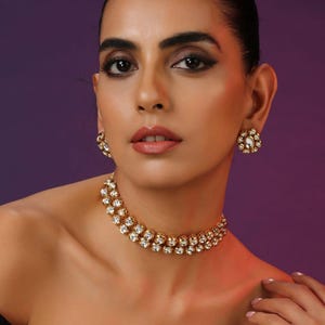 May include: A gold-toned necklace and matching earrings set with clear, round gemstones. The necklace is a choker style, and the earrings are round studs. The jewelry is worn by a person with dark hair and makeup, against a purple background.