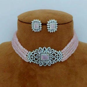 Könnte beinhalten: Eine rosafarbene Perlenkette mit einem silbernen Anhänger aus klarem Kristall. Der Anhänger ist quadratisch mit einem floralen Design. Die Kette wird mit einem Paar passender Ohrringe geliefert.