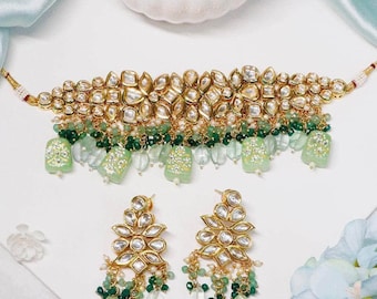 Op de afbeelding: Een goudkleurige ketting en bijpassende oorbellen set. De ketting heeft een bloemenontwerp met heldere en groene accenten, en hangende groene en heldere kralen. De oorbellen hebben een vergelijkbaar ontwerp met hangende kralen. De sieradenset wordt op een wit oppervlak gepresenteerd.