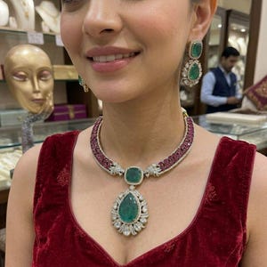 May include: A woman wearing a luxurious jewelry set. The set includes a necklace with a large emerald pendant, ruby accents, and diamond detailing, along with matching emerald and diamond earrings. The jewelry is set against a red velvet top.