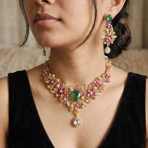May include: A gold-toned necklace and matching drop earring set featuring intricate leaf-shaped metalwork, small pink floral accents, tiny white beads, and prominent teardrop-shaped green stones as the central focal points of the jewelry.