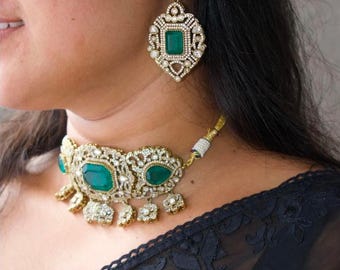 May include: An ornate gold-toned jewelry set featuring a choker necklace and matching earrings. The necklace and earrings are adorned with emerald-green gemstones and clear crystals. The earrings have a large, square emerald-green stone in the center.