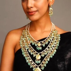 Peut inclure: Un ensemble de collier et boucles d'oreilles assorties. Le collier présente des rangées de perles blanches, des accents dorés et des pierres en forme de goutte d'eau vert clair. Les boucles d'oreilles ont un design similaire, avec des éléments dorés et verts.