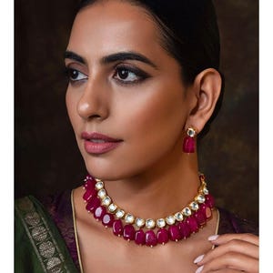 Puede incluir: Un collar de oro con un diseño en capas que presenta piedras rosas y blancas. El collar lo lleva una persona con el pelo oscuro y maquillaje.