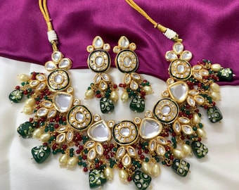 May include: A gold-toned necklace and earring set with clear, round, and emerald-green accents. The necklace and earrings feature a floral design with pearl-like beads and red accents. The jewelry set is displayed on a white and purple background.