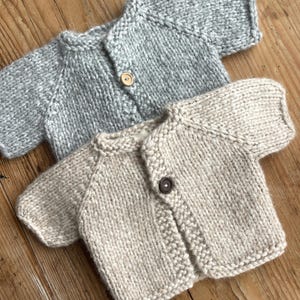 Może przedstawiać: Dwa ręcznie robione sweterki dla niemowląt. Jeden jest jasnoszary, drugi beżowy. Każdy ma krótkie rękawy i pojedyncze zapięcie na guzik. Sweterki wykonane są z grubej przędzy, idealne do ogrzania dziecka.