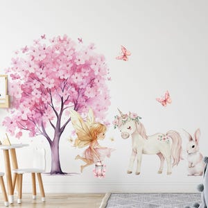 Op de afbeelding: Een roze en witte eenhoorn met een bloemenkrans staat naast een fee met vleugels en een lantaarn. Een roze konijn en vlinders zijn ook inbegrepen in deze fantasierijke wanddecoratieset.