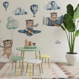 May include: A set of wall decals featuring watercolor-style brown bears in aviator gear flying blue airplanes and hot air balloons, accompanied by blue clouds and balloons. The scene is displayed on a wall above a small round table and three stools.