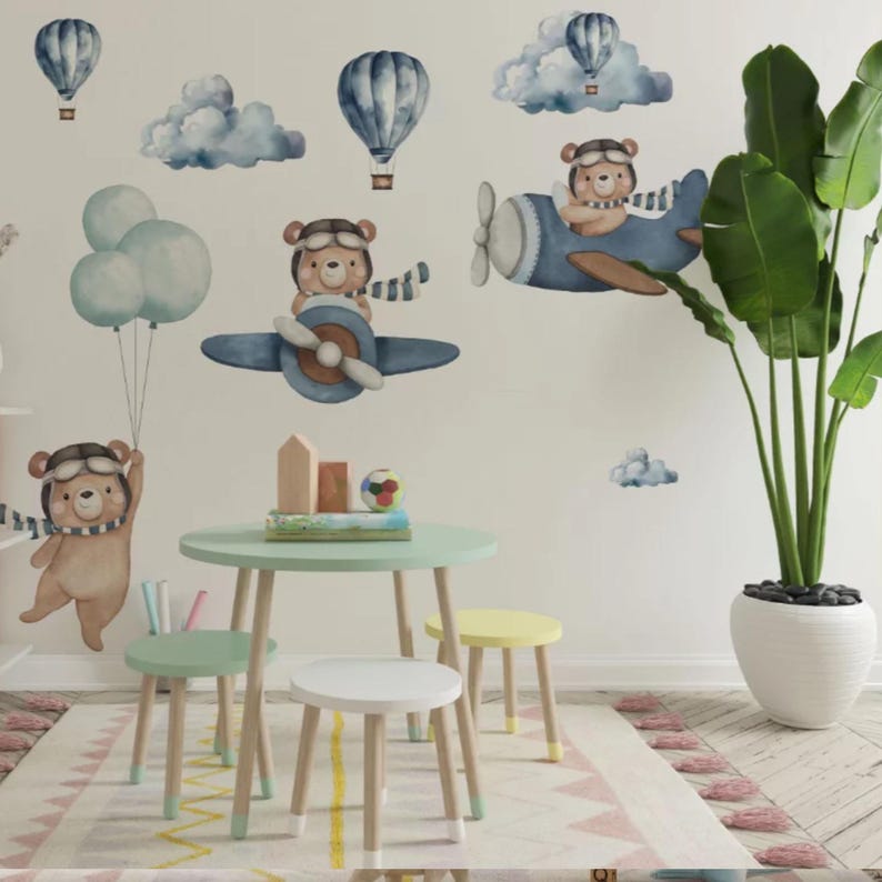 May include: A set of wall decals featuring watercolor-style brown bears in aviator gear flying blue airplanes and hot air balloons, accompanied by blue clouds and balloons. The scene is displayed on a wall above a small round table and three stools.
