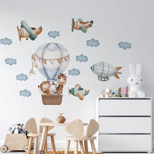 May include: A set of wall decals featuring a watercolor-style hot air balloon carrying a lion, zebra, and giraffe in a brown basket, accompanied by two airplanes, a blimp, and several blue clouds arranged on a plain white wall surface.