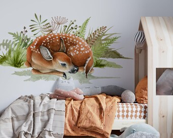 以下が含まれることがあります： 緑のシダと葉のベッドで眠る子鹿の水彩画。子鹿は白点のある茶色で、頭を体に押し付けています。イラストは、茶色と白の毛布とグレーと白の枕のあるベッドの上の白い壁にあります。
