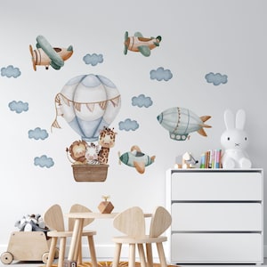 Könnte beinhalten: Ein Set Wandaufkleber mit einem Heißluftballon im Aquarellstil, der einen Löwen, ein Zebra und eine Giraffe in einem braunen Korb trägt, ergänzt durch zwei Flugzeuge, ein Luftschiff und mehrere blaue Wolken auf einer weißen Wandfläche.