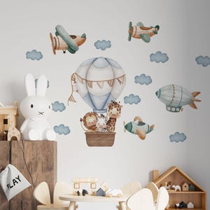 May include: A wall decal set featuring a large hot air balloon carrying a lion, zebra, and giraffe in a woven basket, surrounded by two airplanes, a blimp, and several blue clouds, all rendered in a soft, watercolor-style illustration.