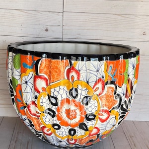 May include: A large, round ceramic planter featuring a vibrant, hand-painted floral pattern in shades of orange, yellow, red, and green against a white background. The bowl-shaped vessel has a scalloped rim with a solid black painted edge.