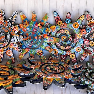 May include: A collection of seven sun-shaped ceramic wall plaques featuring intricate, multi-colored floral and geometric patterns. Each piece has a central black spiral design surrounded by radiating points painted in vibrant hues of orange, blue, and red.