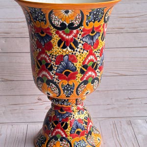 May include: A tall, ornate ceramic vase with a wide, flaring rim and a pedestal base. The vase is painted with a vibrant floral pattern in red, blue, yellow, and orange against a black and white background. The rim and base are orange.