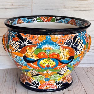 May include: A large, round ceramic planter featuring a vibrant, hand-painted floral pattern in shades of orange, blue, yellow, green, and black. The bowl-shaped vessel has a wide, dark blue rim and a pedestal base, with intricate, colorful botanical motifs covering the entire exterior surface.