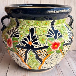 May include: A large, round ceramic planter with a wide, dark blue rim and two handles. The planter features a colorful floral design with green, orange, red, and black accents. The design includes stylized flowers and geometric patterns.