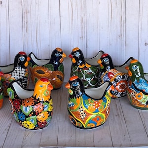 May include: A collection of colorful ceramic hen-shaped planters. Each planter features a unique design with vibrant floral patterns, geometric shapes, and bold color combinations. The hens have open tops for planting and are displayed on a light wood surface.
