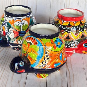May include: Three ceramic turtle planters with vibrant, hand-painted designs. Each planter features a dark blue turtle head and feet, with colorful floral patterns in orange, green, and blue. The rims are dark blue or red.