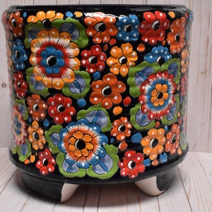 May include: A black ceramic planter with a floral design. The planter has a round shape and three feet. The flowers are painted in shades of red, orange, yellow, blue, and green.