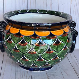 May include: A ceramic planter with a vibrant, hand-painted design. The planter features a scalloped pattern in green, orange, and red, with a dark blue rim and handles. The interior is white.