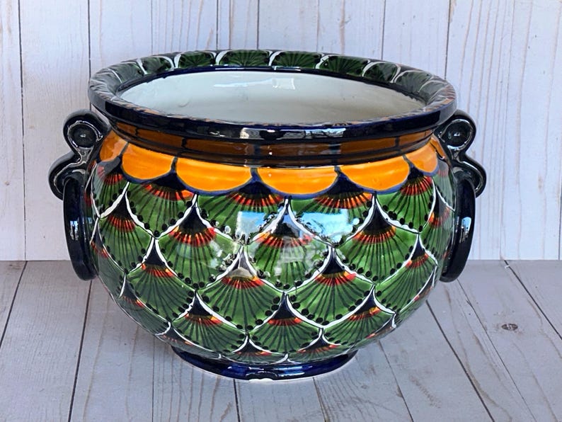 May include: A ceramic planter with a vibrant, hand-painted design. The planter features a scalloped pattern in green, orange, and red, with a dark blue rim and handles. The interior is white.