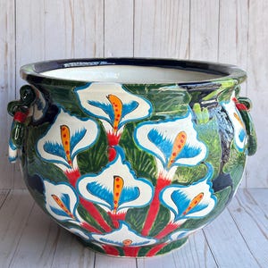 May include: A large, round ceramic planter with a hand-painted floral design. The planter features white calla lilies with blue and orange accents against a green background. The rim is dark blue, and it has two handles. The planter is on a light wood surface.