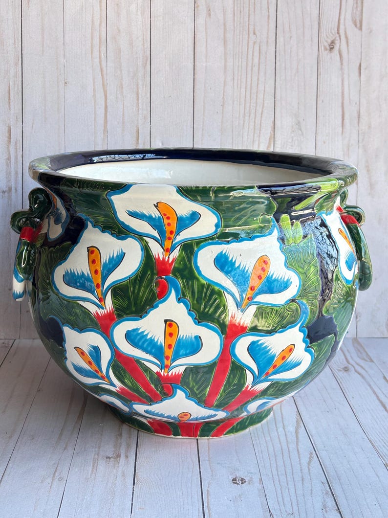 May include: A large, round ceramic planter with a hand-painted floral design. The planter features white calla lilies with blue and orange accents against a green background. The rim is dark blue, and it has two handles. The planter is on a light wood surface.