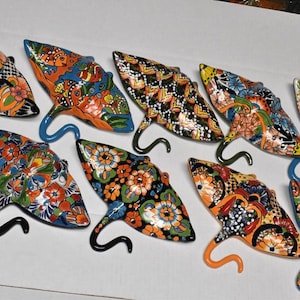 May include: A collection of nine ceramic stingray wall decorations, each featuring vibrant, hand-painted floral and geometric patterns in various color combinations including orange, blue, green, black, and white, with a curved tail hook.