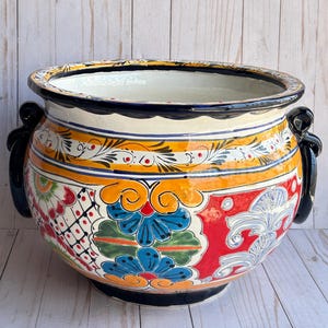 May include: A large, round, hand-painted ceramic planter with a wide rim and two black handles. The planter features a vibrant design with blue, orange, red, and white floral patterns. The interior is white.