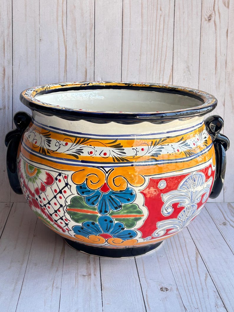 May include: A large, round, hand-painted ceramic planter with a wide rim and two black handles. The planter features a vibrant design with blue, orange, red, and white floral patterns. The interior is white.
