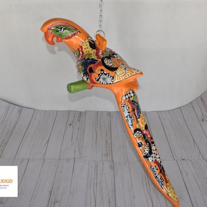May include: A vibrant, hand-painted ceramic parrot sculpture in shades of orange, green, and blue. The parrot is suspended from a chain and features intricate floral and geometric patterns. The sculpture has a green perch and is a decorative piece.