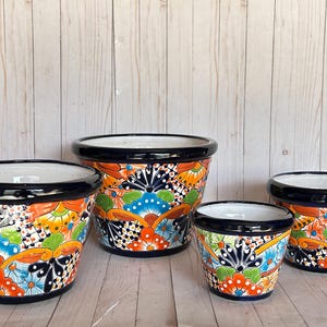 May include: A set of four ceramic planters featuring a vibrant Talavera-style pattern with orange, blue, green, and black floral and geometric motifs. Each pot has a glossy white interior and a thick, solid black rim around the top edge.