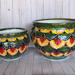 May include: Two ceramic planters with a vibrant, hand-painted design. The planters feature a dark green base, with a repeating pattern of yellow, red, and white accents. Each planter has a white interior and green handles.