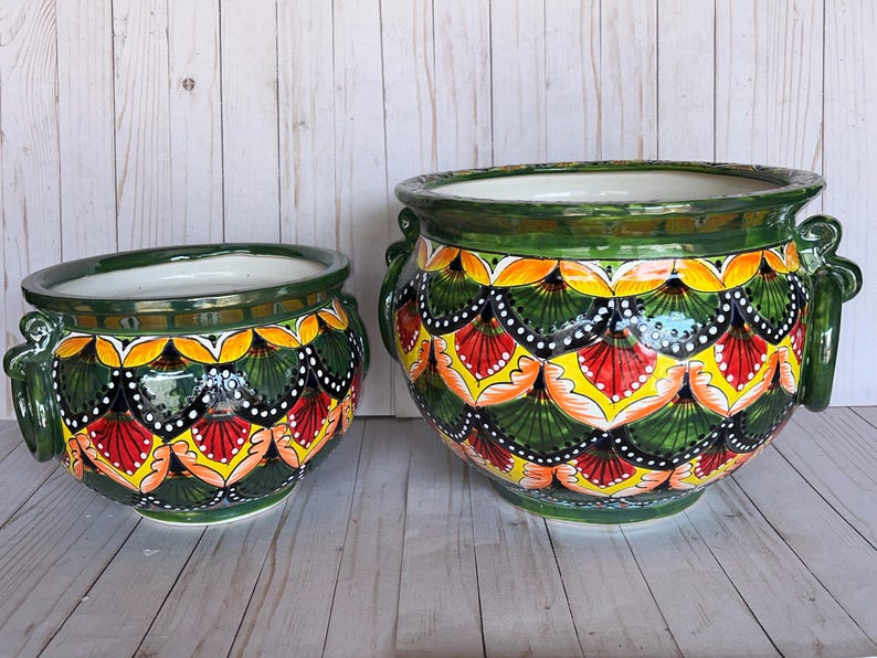 May include: Two ceramic planters with a vibrant, hand-painted design. The planters feature a dark green base, with a repeating pattern of yellow, red, and white accents. Each planter has a white interior and green handles.