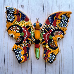 May include: A vibrant, hand-painted ceramic butterfly with intricate floral patterns in red, yellow, blue, and black. The butterfly has a bright orange body and wings, and is a decorative wall hanging.
