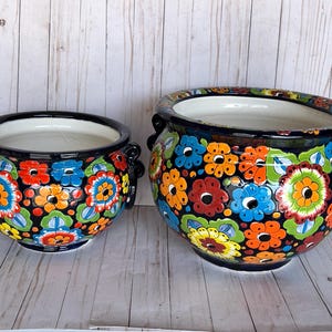 May include: Two ceramic flower pots with a black base and colorful floral designs. The pots have a white interior and black handles. The larger pot is approximately 12 inches in diameter, and the smaller pot is about 8 inches in diameter.