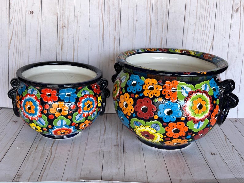 May include: Two ceramic flower pots with a black base and colorful floral designs. The pots have a white interior and black handles. The larger pot is approximately 12 inches in diameter, and the smaller pot is about 8 inches in diameter.