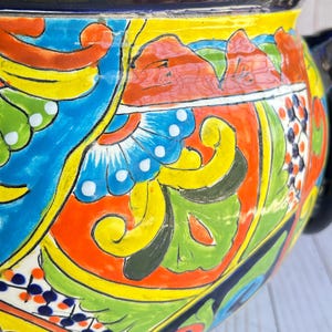 May include: A vibrant, hand-painted ceramic pot with a black rim and handle. The pot features a colorful design with blue, yellow, orange, and green floral and geometric patterns. The design includes stylized flowers and leaves, creating a decorative piece.