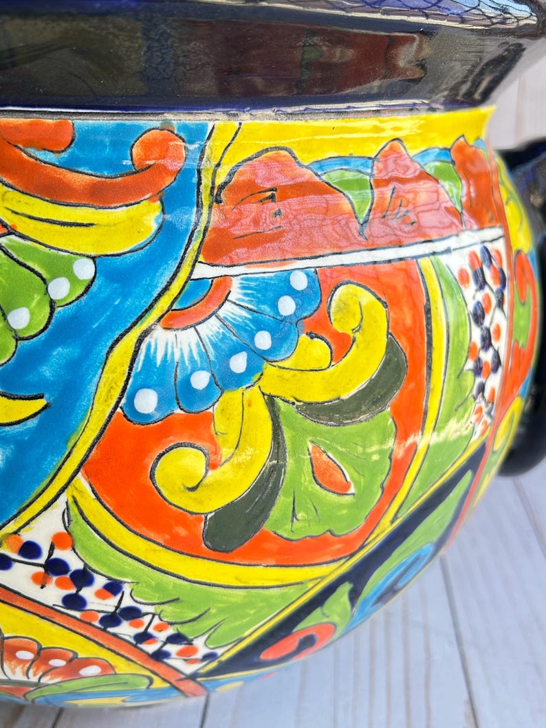May include: A vibrant, hand-painted ceramic pot with a black rim and handle. The pot features a colorful design with blue, yellow, orange, and green floral and geometric patterns. The design includes stylized flowers and leaves, creating a decorative piece.