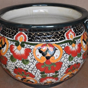 May include: A round, ceramic planter with a wide opening and two side handles. The planter features a hand-painted floral design in red, orange, and green on a white background. The rim and handles are dark green.