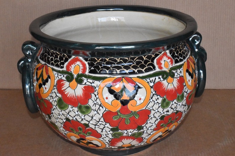 May include: A round, ceramic planter with a wide opening and two side handles. The planter features a hand-painted floral design in red, orange, and green on a white background. The rim and handles are dark green.