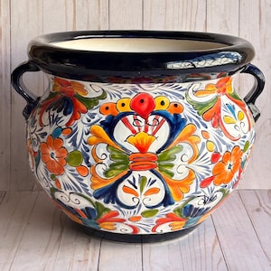 May include: A large, round ceramic planter with a vibrant floral design. The planter features a white base with colorful flowers in orange, blue, and green. It has a black rim and handles, and is suitable for indoor or outdoor use.