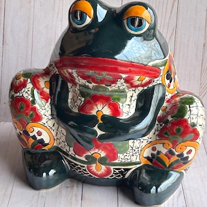 May include: A ceramic frog figurine with a glossy, dark green finish. The frog's eyes are blue with orange accents, and its mouth is red. The frog's body is decorated with red and yellow floral patterns on a white background.